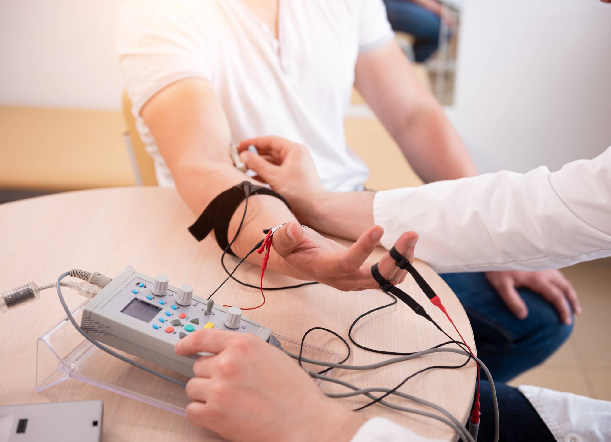 A person receives a medical test on their arm using electrodes connected to a diagnostic machine, while a healthcare professional operates the device.