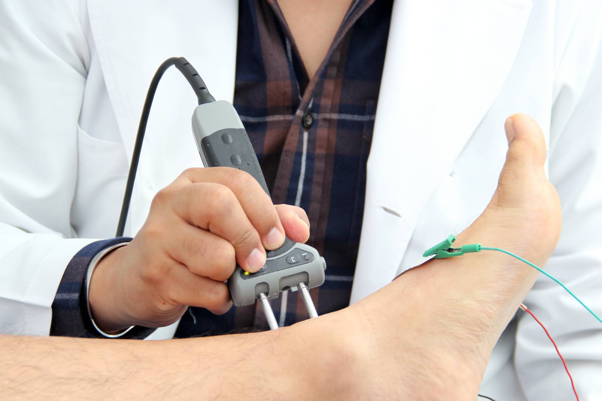 A doctor wearing a white coat holds a medical device against a patient's ankle for a nerve conduction test; wires and electrodes are attached to the foot.