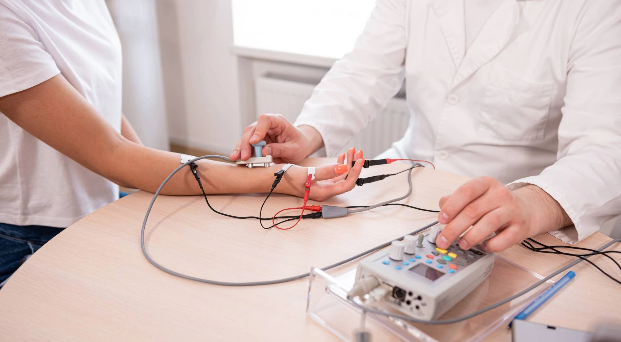 A healthcare professional attaches electrodes to a patient's fingers and wrist while operating a medical device on a table, possibly for a nerve conduction or electrophysiology test.